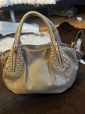 Beige handväska med nitar - Beige/ ljusbrun handväska med guldiga nitar och diamanter. 28 cm på längden 41 cm på bredden. Seg att öppna där det är en mörk fläck på bilden på dragkedjan, skriv för mer frågor☺️☺️