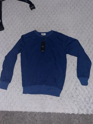 Blå sweatshirt från Stone Island - Blå sweatshirt från Stone Island med rund halsringning och ribbade muddar vid ärmslut och midja. Klassisk och stilren design med diskret logga på ärmen. Perfekt för en avslappnad och trendig look. Sido plagget är tyvärr inte med, men det finns och köpa ganska billigt!!!! Aldrig använt!