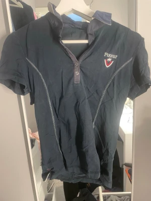 Mörkblå Pikeur t-shirt med detaljer - Snygg mörkblå t-shirt från Pikeur med korta ärmar och kontrastkrage. Framsidan har Pikeur-logga och emblem, baksidan har stor broderad text. Snygga sömdetaljer och axelpartier med ränder ger en sportig vibe. Perfekt för dig som gillar stilrena och sportiga plagg.