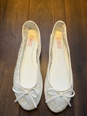 Vita spetsballerinas från Nelly - Snygga vita ballerinas från Nelly med spetsdetaljer och en söt rosett framtill. Skorna har en rund tå och är tillverkade i textil med spetsmönster. Perfekta för dig som gillar en feminin och stilren look.