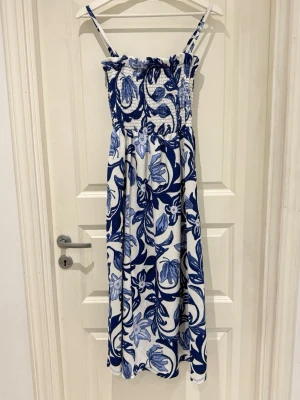 Blåvit blommig långklänning - Snygg långklänning med smala axelband och smockad överdel. Klänningen är vit med blått blommönster och har en luftig passform, perfekt för varma dagar. Tillverkad i ett mjukt och lätt material som känns skönt mot huden.