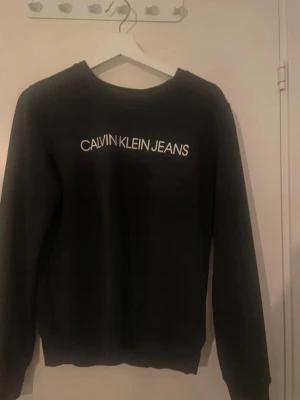 Svart sweatshirt från Calvin Klein Jeans - Svart sweatshirt med vit Calvin Klein Jeans-logga tryckt på bröstet. Klassisk rund halsringning och långa ärmar. Perfekt för en avslappnad och stilren look.