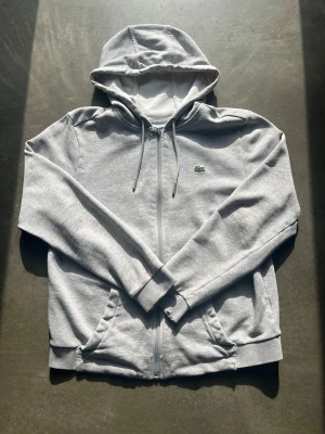 Grå zip-hoodie från Lacoste - Snygg grå hoodie med dragkedja från Lacoste. Klassisk krokodillogga på bröstet, justerbar huva med snören och två fickor framtill. Använd och sliten vid ärmarna och längst ner vid magen men fortfarande snygg. Hör av er vid frågor pris kan diskuteras!😁