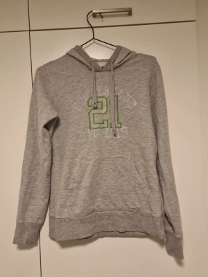 Grå hoodie med grön 21-tryck - Grå hoodie med huva och dragsko, samt en stor ficka framtill. På bröstet finns ett tryck med siffran 21 i grönt och vitt. Hoodien har långa ärmar och är tillverkad i mjukt sweatshirtmaterial, perfekt för en avslappnad stil.