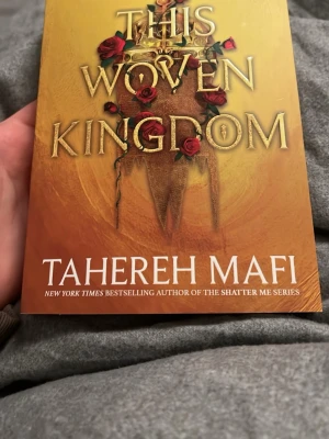 This Woven Kingdom av Tahereh Mafi - En fantasyroman på engelska med gyllene omslag och röda rosor, skriven av Tahereh Mafi. Boken handlar om Alizeh, en tjänare med hemlig bakgrund, och prins Kamran. Perfekt för dig som gillar episka berättelser om magi, kärlek och maktkamp.