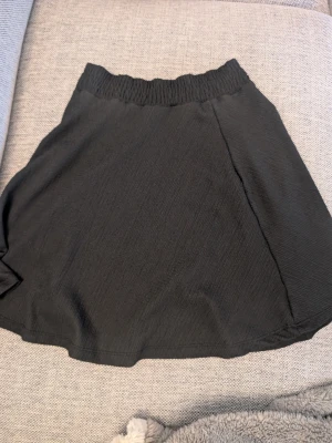 Svart kortkjol med resårmidja stl xs s - En svart kortkjol med bred resår i midjan och ett lätt strukturerat tyg. Kjolen har en enkel och stilren design som passar till många olika looks. Perfekt för dig som gillar minimalistisk stil.