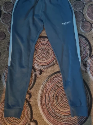 Blå mjukisbyxor från McKenzie - Snygga blå mjukisbyxor från McKenzie med vita sidoremsor och resår i midjan. Byxorna har smal passform och muddar vid bensluten. Perfekta för en avslappnad och sportig stil.