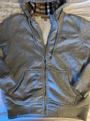 Burberry hoodie grå - Snygg grå hoodie med dragkedja från Burberry. Klassisk rutmönstrad huva, broderad logga på bröstet och stora fickor framtill. Mjuk bomullsblandning och ribbade muddar. Perfekt för en chill och stilren look. Skriv om ni har frågor. Pris går att diskuteras.