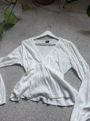 Vit omlottblus med v-ringning - Snygg vit omlottblus från gina tricot med djup v-ringning och långa, vida ärmar. Blusen har en lätt rynkad midja och är tillverkad i ett tunt, krinklat tyg som ger en luftig känsla. Perfekt att styla med jeans eller kjol för en trendig look.