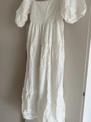 Vit långklänning med puffärmar - Romantisk vit långklänning med broderade detaljer och puffiga kortärmar. Klänningen har smock upptill och är tillverkad i bomull, vilket gör den luftig och perfekt för varma dagar. Snygg och trendig modell med voluminös kjol.