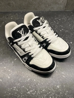 Louis Vuitton svartvita sneakers - Svartvita Louis Vuitton sneakers med klassisk LV-logga på plösen och präglad monogramdetalj på lädret. Skorna har vita snören, svart och vit ovandel i läder och textil samt robust sula. Snygg kontrast mellan färgerna och exklusiv känsla.