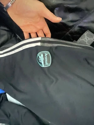 Arsenal x Adidas svarta träningsbyxor - Svarta träningsbyxor från Adidas i samarbete med Arsenal. Byxorna har vita ränder längs sidorna, Arsenal-logga och Adidas-logga i ljusblått. Perfekta för fotbollsträning eller chill. Materialet är troligtvis polyester för sportig känsla.