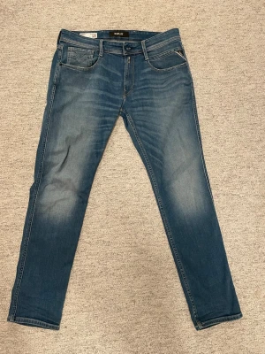 Blå slim fit jeans från Replay - Snygga blå jeans från Replay i modellen Anbass med slim fit passform. Jeansen har klassisk femficksdesign, diskreta slitningar och ljusare partier på låren för en cool look. Tillverkade i mjukt denimtyg som sitter skönt hela dagen.