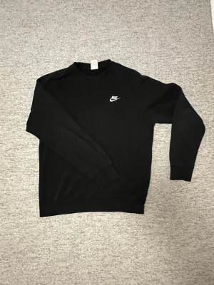 Svart sweatshirt från Nike - Svart sweatshirt från Nike med klassisk logga broderad på bröstet. Tröjan har rund halsringning och ribbade muddar vid ärmslut och nederkant. Perfekt för en avslappnad och sportig look.
