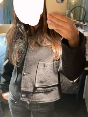  teddyjacka  - Cool svart teddyjacka med stora framfickor och knappar framtill. Jackan har en croppad passform och är perfekt för dig som gillar en avslappnad och trendig stil. Materialet är mjukt och ger en skön känsla. Långärmad och enkel att matcha med jeans eller kjol.