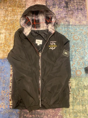 Svart Burberry vindjacka med huva- unik - Svart vindjacka från Burberry med huva och klassiskt rutigt foder. Jackan har dragkedja framtill, logotyp på bröstet och detaljer på ärmen. Materialet är lätt och quiltat, perfekt för att hålla vinden ute. Snygga Burberry-tryck längs sidorna och modern passform.