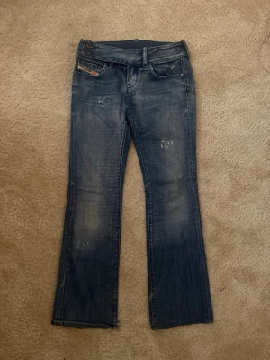 Blå lågmidjade utsvängda jeans från Diesel - Säljer ett par blå bootcut jeans från Diesel med slitna detaljer och dubbla knappar i midjan. Midjemått 36cm 💕