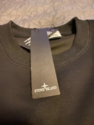Svart sweatshirt från Stone Island - Säljer en svart sweatshirt från Stone Island med klassisk rund halsringning och ribbade muddar. Tröjan har en stilren look och är tillverkad i mjukt bomullsmaterial. Perfekt för dig som gillar streetwear och vill ha något enkelt men ändå exklusivt.