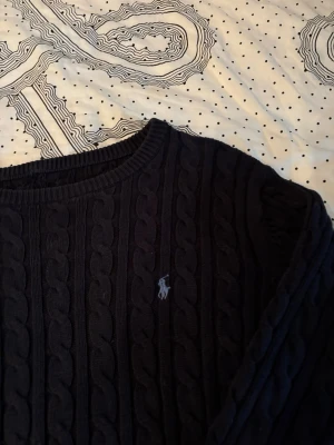 Mörkblå kabelstickad tröja Polo Ralph Lauren - Kabelstickad mörkblå tröja från Polo Ralph Lauren med rund halsringning och den klassiska broderade loggan på bröstet. Tröjan har långa ärmar och ribbade muddar, perfekt för lager-på-lager och en stilren look.