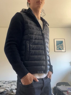 Svart pufferjacka med stickade ärmar - Säljer en svart pufferjacka med stickade ärmar och huva. Jackan har quiltad front och rygg i glansigt material, dragkedja framtill och två fickor med dragkedja. Perfekt för kalla vinterdagar och riktigt snygg streetstil.