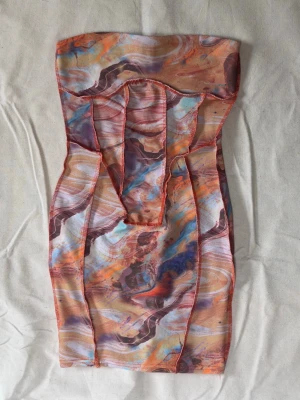 Färgglad tubklänning med mönster - Säljer en kort, ärmlös tubklänning med färgstarkt abstrakt mönster i orange, blått, lila och beige. Klänningen har en figurnära passform och är sydd i ett stretchigt syntetmaterial. Perfekt för dig som gillar unika plagg med mycket färg.