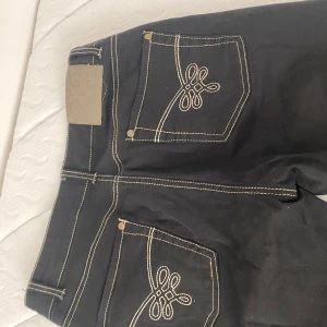 Mid waist Boatcut jeans från Minipris - Snygga svarta jeansbyxor från Minipris med dekorativa sömmar på bakfickorna. Byxorna har en klassisk design med knapp och dragkedja. Fint mönster på bakficka