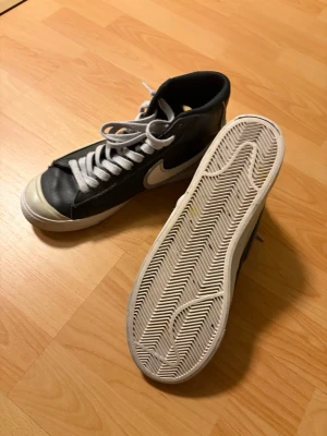 Nike svartvita high-top sneakers - Säljer ett par klassiska high-top sneakers från Nike i svart och vitt. Skorna har vit sula, vit tå och vita skosnören samt den ikoniska swoosh-loggan på sidan. Materialet är läder och gummi, perfekt för dig som vill ha en cool och tidlös look.