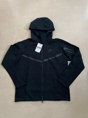 Nike tech fleece zip hoodie svart storlek large - Svart hoodie från Nike med hel dragkedja och huva. Tröjan har diskreta svarta detaljer över bröstet och Nike-logga på vänster sida. Tillverkad i mjukt material som känns skönt mot huden. Perfekt för en sportig och avslappnad stil.