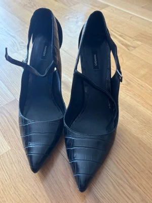 Svarta pumps med krokopräglat mönster Mango - Snygga svarta pumps från Mango med spetsig tå och krokopräglat mönster. Skorna har smal klack, öppen häl och justerbart spänne vid ankeln. Tillverkade i konstläder med glansig finish för en trendig look.