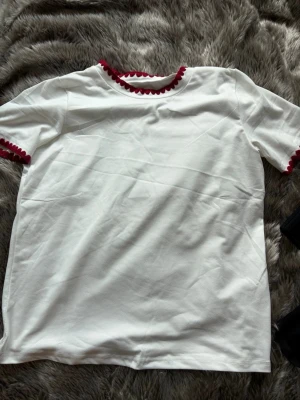 Vit t-shirt med röda volangdetaljer - Vit t-shirt med korta ärmar och röda volangdetaljer vid halsringning och ärmslut. 