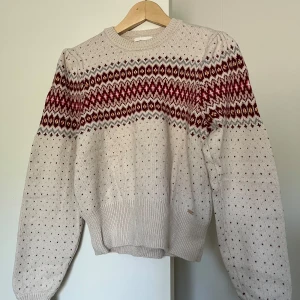 Beige stickad tröja med mönster - Stickad tröja i beige med klassiskt mönster i vinrött, brunt och vitt över bröstet och axlarna. Tröjan har rund hals, långa ärmar och ribbstickade muddar. Perfekt för kalla dagar och har en mysig, retro känsla. Använd få gånger. 