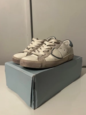 Vita Philippe Model sneakers med grå detaljer - Snygga vita sneakers från Philippe Model med grå mockadetaljer och blått parti vid hälen. Dom är använda lite men ändå i väldigt bra skick. Skorna är storlek 40.