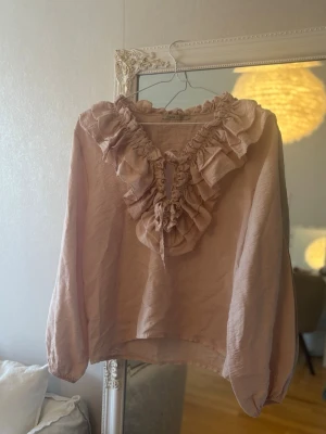 Ljusrosa blus med volangdetaljer - Supersöt ljusrosa blus med långa ärmar och volangdetaljer längs v-ringningen. Blusen har en lös passform och är tillverkad i ett lätt, skrynkligt tyg som ger en avslappnad känsla. Perfekt för dig som gillar romantiska detaljer.