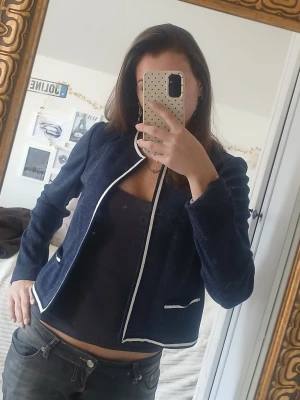 Mörkblå kofta med vita detaljer - Snygg mörkblå kofta från M&S med vita kontrastkanter längs krage, öppning och fickor. Rak, öppen modell utan knappar och med långa ärmar. Perfekt för en stilren look och enkel att matcha med olika outfits.