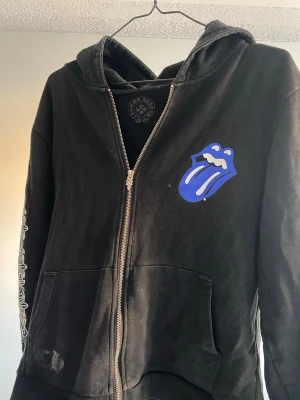 Svart hoodie Chrome Hearts x Rolling Stones - Svart hoodie med dragkedja från Chrome Hearts x Rolling Stones. Stor blå Rolling Stones-tunga på bröstet och ryggen, vita tryck på ärmen och rygg, samt fickor framtill. Något solblek, därav priset.