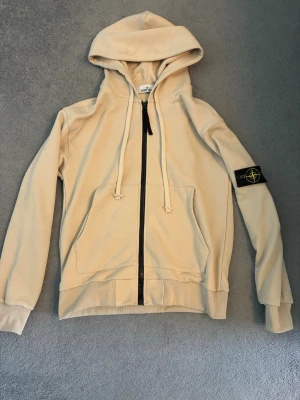 Beige hoodie från Stone Island - Säljer en beige hoodie från Stone Island med dragkedja framtill, stor huva och känguruficka. Klassisk svart Stone Island-patch på vänster ärm. Tillverkad i mjuk bomull, perfekt för en chill och stilren look.