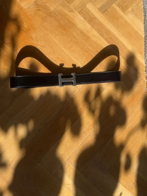 Svart Herms bälte med H-spänne - Snyggt svart bälte från Herms med det ikoniska H-spännet i silverfärgad metall. Bältet är gjort i slätt skinn och passar perfekt till jeans eller kostymbyxor. En klassisk accessoar som lyfter vilken outfit som helst.