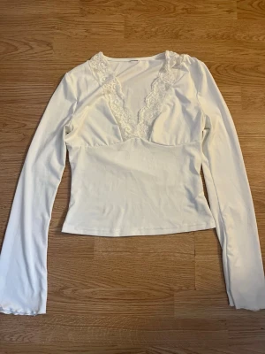 Vit långärmad topp med spetsdetalj - Snygg vit långärmad topp med djup v-ringning och spetsdetaljer längs urringningen. Figurnära passform💕skriv vid frågor 💕
