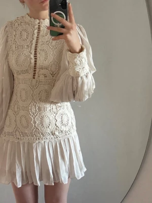 Vit spetsklänning med volang studentklänning  - Romantisk vit kortklänning med spetsdetaljer och volangkant. Klänningen har långärmade, lätt transparenta ärmar och vackert broderat mönster framtill och baktill. Hög halsringning och dragkedja i ryggen. Perfekt för dig som gillar vintageinspirerad stil. Virkad, spets, vintage, ballongärmar perfekt till studenten! Prislapp är med!