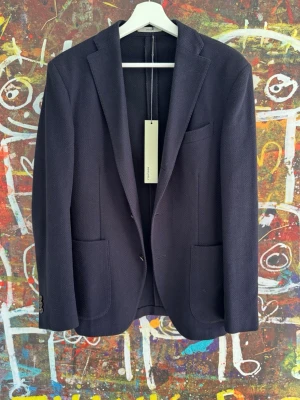 Mörkblå kavaj från Boglioli - Stilren mörkblå kavaj från Boglioli med klassisk passform och diskret fiskbensmönster. Kavajen har två framfickor, bröstficka och knappar vid ärmslut. Tillverkad i ett mjukt tyg som ger en exklusiv känsla. Nypris runt 10-12 k 