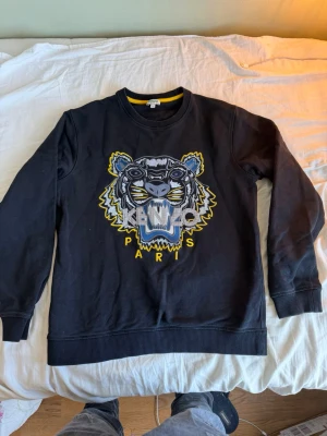 Kenzo sweatshirt - Svart sweatshirt från Kenzo med ikoniskt tigerbroderi och texten 'Kenzo Paris' framtill. Tröjan har rund halsringning med gul detalj på insidan och ribbade muddar vid ärmslut och nederkant. Perfekt för dig som gillar streetwear och statement-plagg.