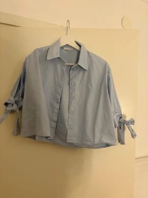 Ljusblå skjorta med knyt från Zara - Trendig ljusblå skjorta från Zara med knytband vid ärmsluten och klassisk krage. Skjortan har knappar framtill och en loose passform som ger en avslappnad vibe. Perfekt för dig som gillar en stilren men ändå unik look.