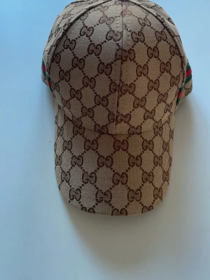 G-keps - Säljer en beige keps med det klassiska GG-monogrammet över hela kepsen. På sidan finns en grön och röd rand som ger extra lyxig känsla. Kepsen är tillverkad i canvasmaterial och har böjd skärm. Perfekt för dig som vill ha en ikonisk accessoar.