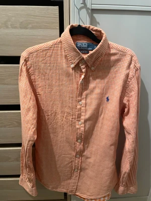 Rutig orange skjorta från Polo Ralph Lauren - Säljer en snygg orange och vit rutig skjorta från Polo Ralph Lauren i slim fit. Skjortan har långa ärmar, klassisk krage och knappar framtill. Ikoniska blå loggan broderad på bröstet. Perfekt för dig som gillar stilrena och fräscha skjortor.