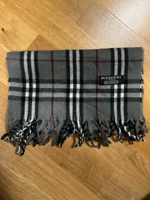 Grå rutig halsduk från Burberry äkta! - Klassisk halsduk från Burberry i grått med svart, vitt och rött rutmönster. Tillverkad i 100% lammull och har tvinnade fransar i ändarna. Märkt med Burberry London-label. Perfekt accessoar för att lyfta din outfit. Såklart äkta tveka aldrig på att att ställa frågor jag svarar snabbt!