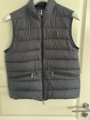 Mörkgrå dunväst från Moncler - Snygg mörkgrå dunväst från Moncler med hög krage och två dragkedjeförsedda fickor framtill. Västen har quiltad design och Moncler-logga på bröstet. Perfekt för lager-på-lager och håller dig varm under kyliga dagar. Riktigt skön till våren, i storleksmått 2 av moncler, typ medium, inköpt på NK i göteborg för 2 år sedan för 8999kr men använd varsamt, skriv gärna ett bud i DM!