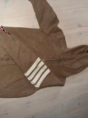 thom browne - Stickad hoodie i beige med dragkedja framtill och fickor. Ena ärmen har vita och beigea ränder vid mudden. På ryggen finns ett stort segelbåtsmotiv i vitt, svart och rött. Tröjan har huva och ribbstickade detaljer. Perfekt för dig som gillar nautisk stil.