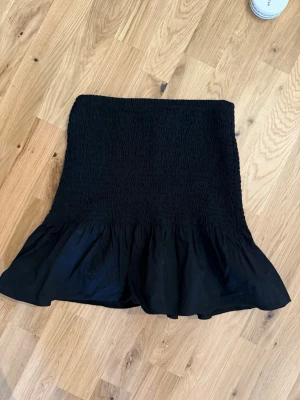 Svart volangkjol med smock - Supersnygg svart kortkjol med smock upptill och bred volang nedtill. Kjolen har en tight passform upptill och faller ut i en volangig kant. Perfekt att styla med en crop top eller stickad tröja. Materialet känns mjukt och stretchigt.