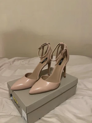 Ljusrosa/nude klackar - Säljer ett par nude-färgade pumps från ONLY med spetsig tå och smal klack. Skorna har glansig finish i syntetmaterial och justerbar ankelrem med spänne för extra support. Perfekta för dig som gillar stilrena och eleganta skor med en modern touch.