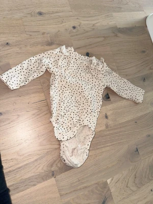 Prickig body med volang från H&M - Söt långärmad body från H&M i ljusbeige med svarta prickar och volangdetaljer vid axlarna. Tillverkad i mjuk bomull och har tryckknappar i grenen för enkel påklädning. Perfekt för en gullig och bekväm vardagslook.